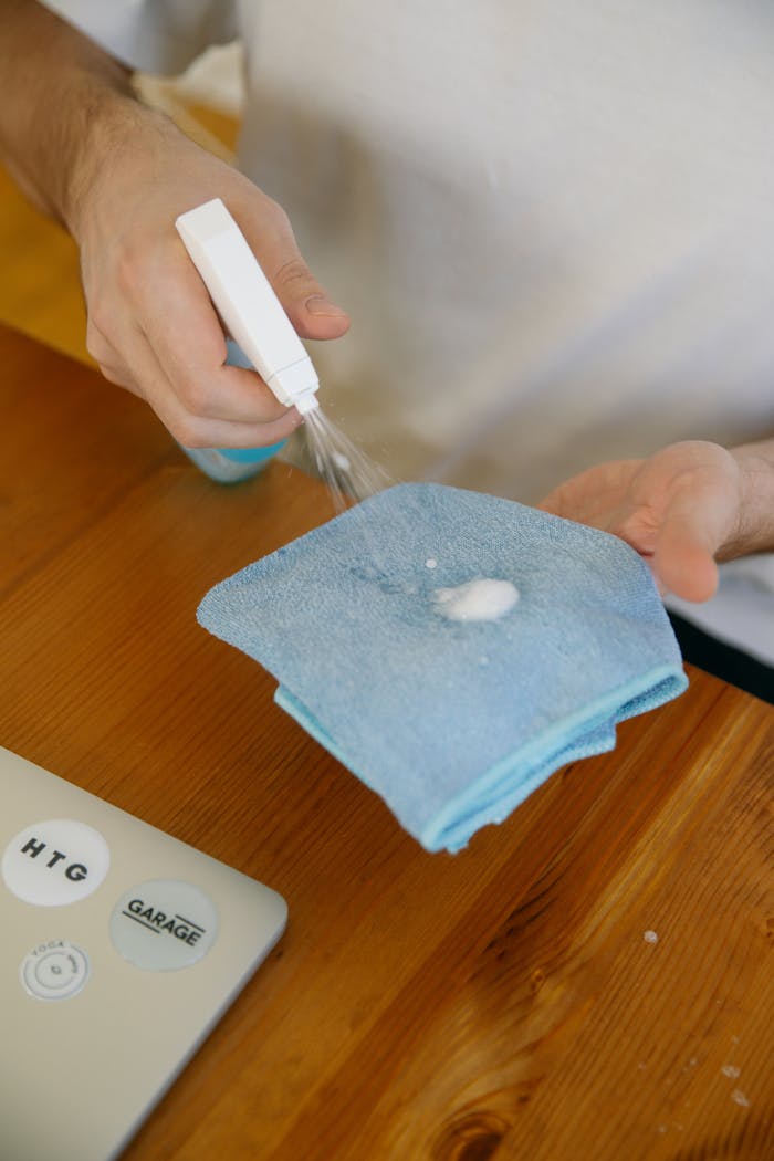 Close-up of hands using disinfectant spray and cloth on a wooden table indoors.